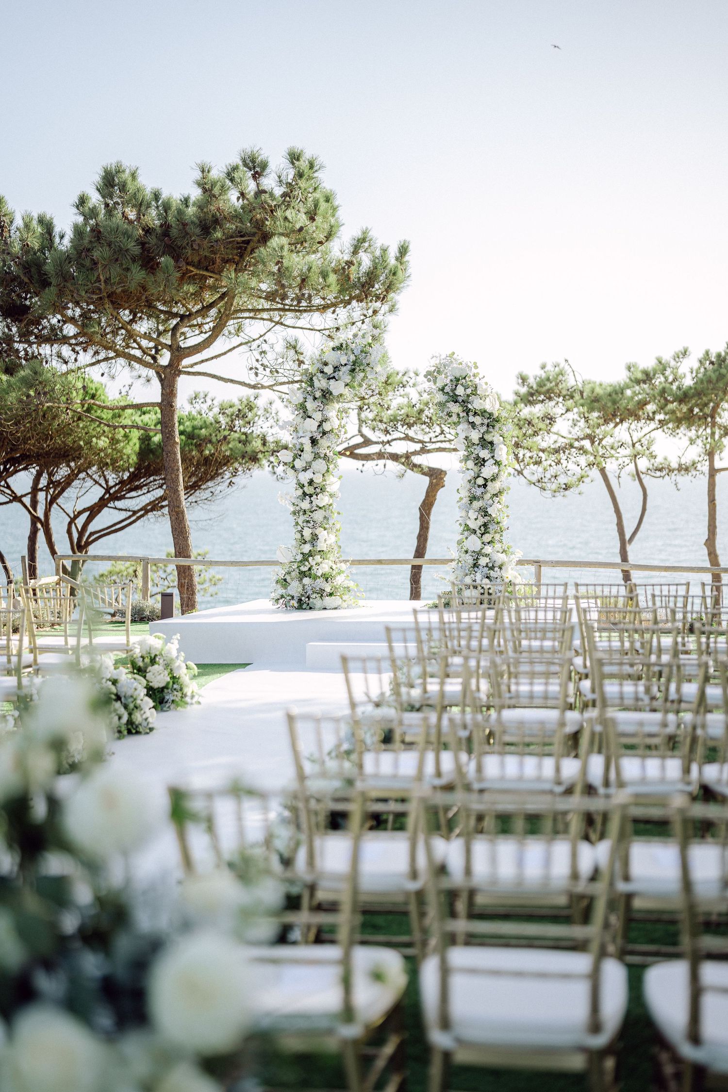 Dreamy wedding ceremony setup with white floral arrangements and gold chairs under pine trees.