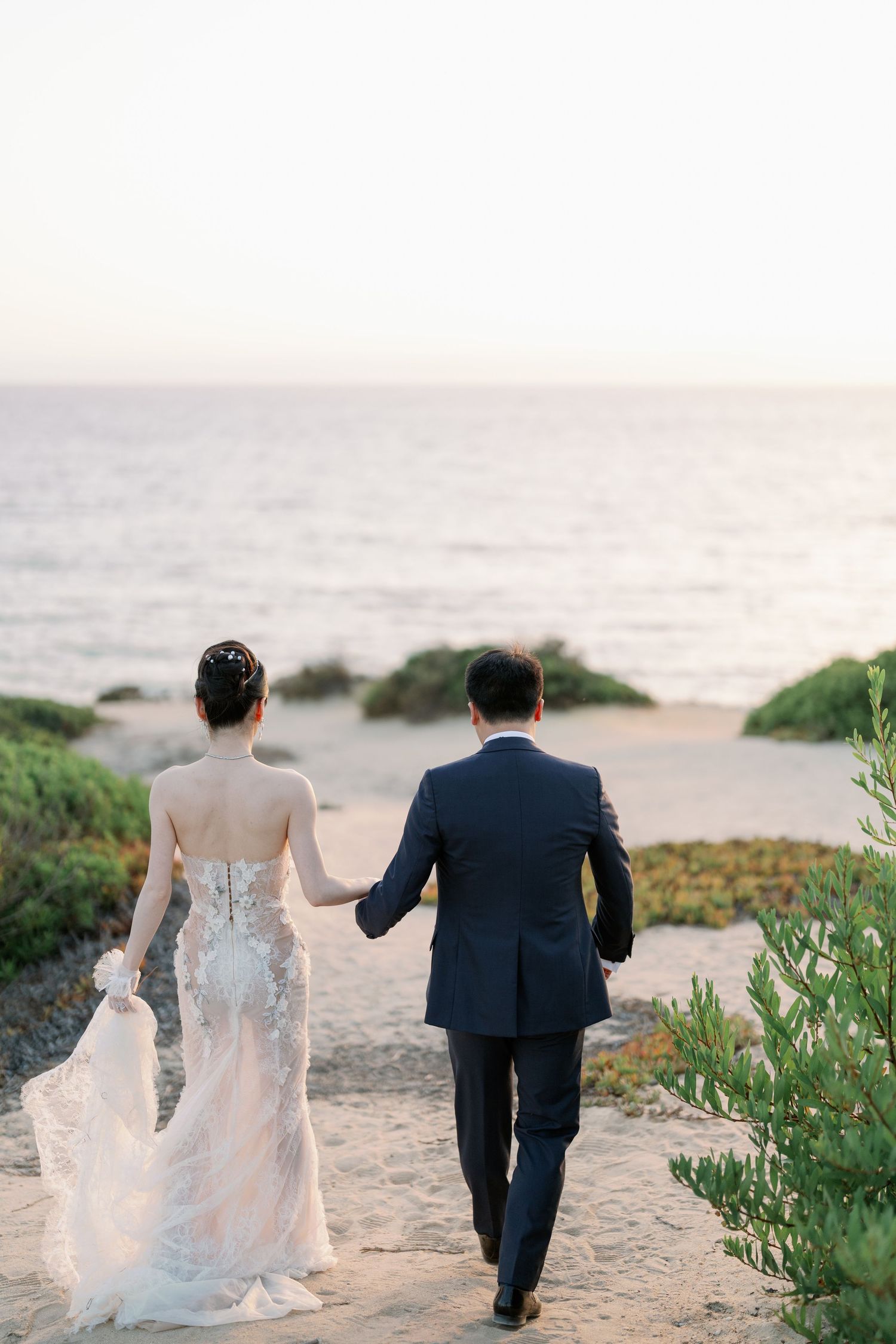 Newlyweds walk hand in hand along coastal path during sunset photo session.