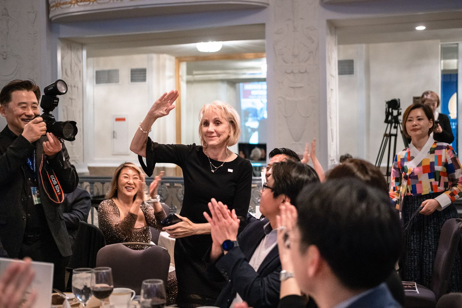 Elegant woman waves to applauding guests at a formal indoor gathering with cameras capturing the celebration.