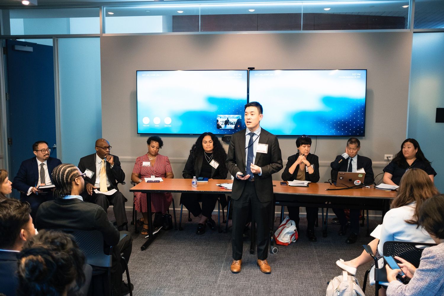 Professional speaker addresses diverse group of business attendees in modern conference room with large display screen.