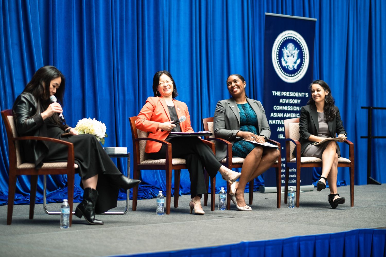 Four women panelists discuss at the President's Advisory Commission on Asian Americans event on a blue-draped stage.
