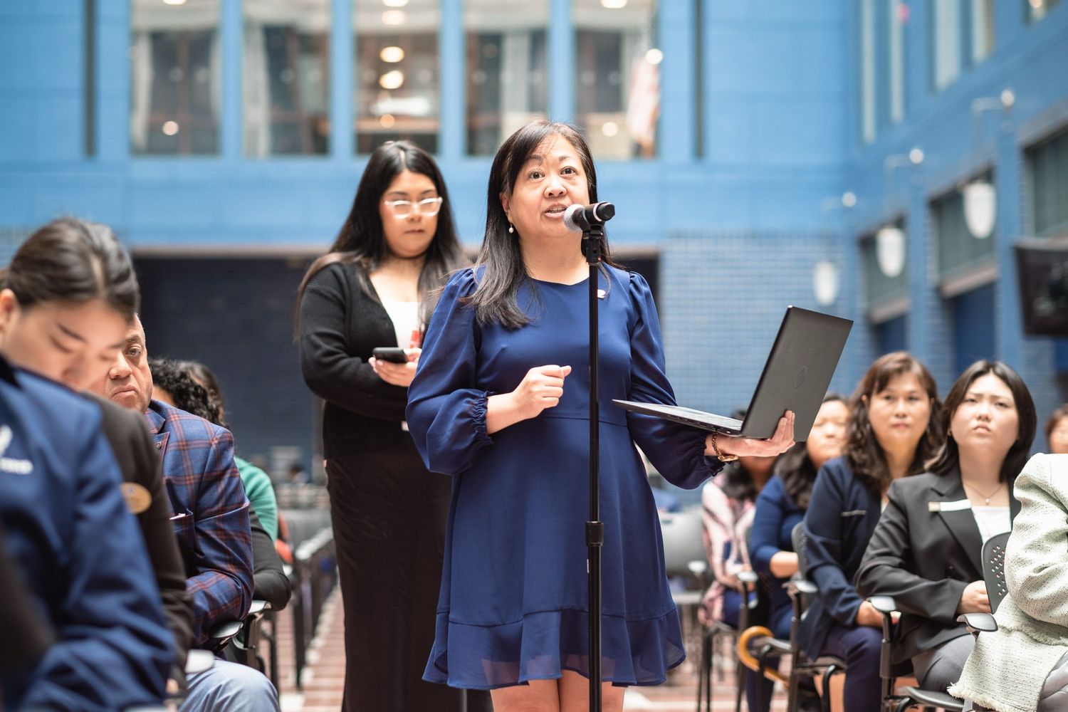 Woman in navy dress speaks at formal event, holding laptop, with seated audience in modern venue.
