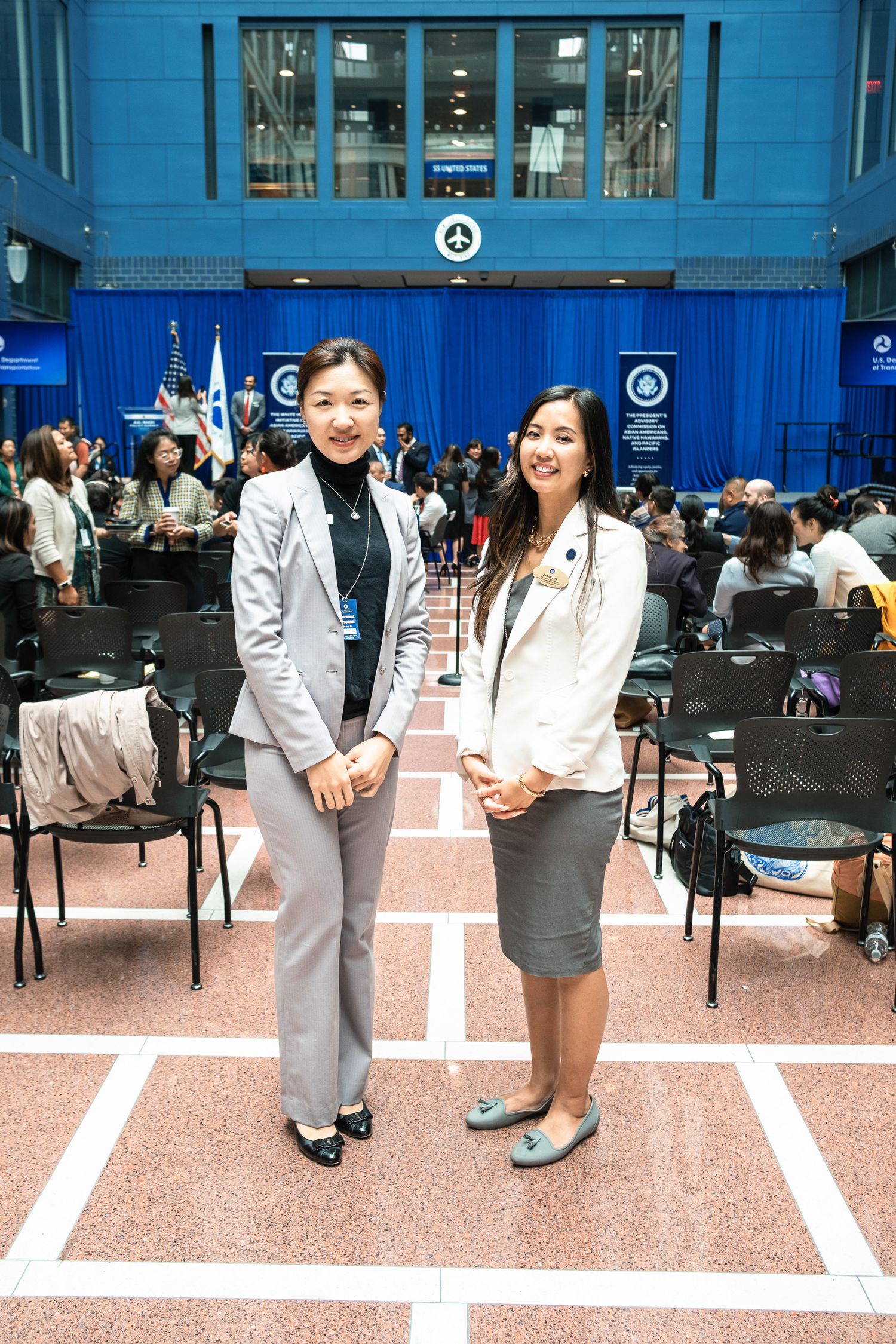 Two women in professional attire smile together at a formal event in a blue-decorated government building.
