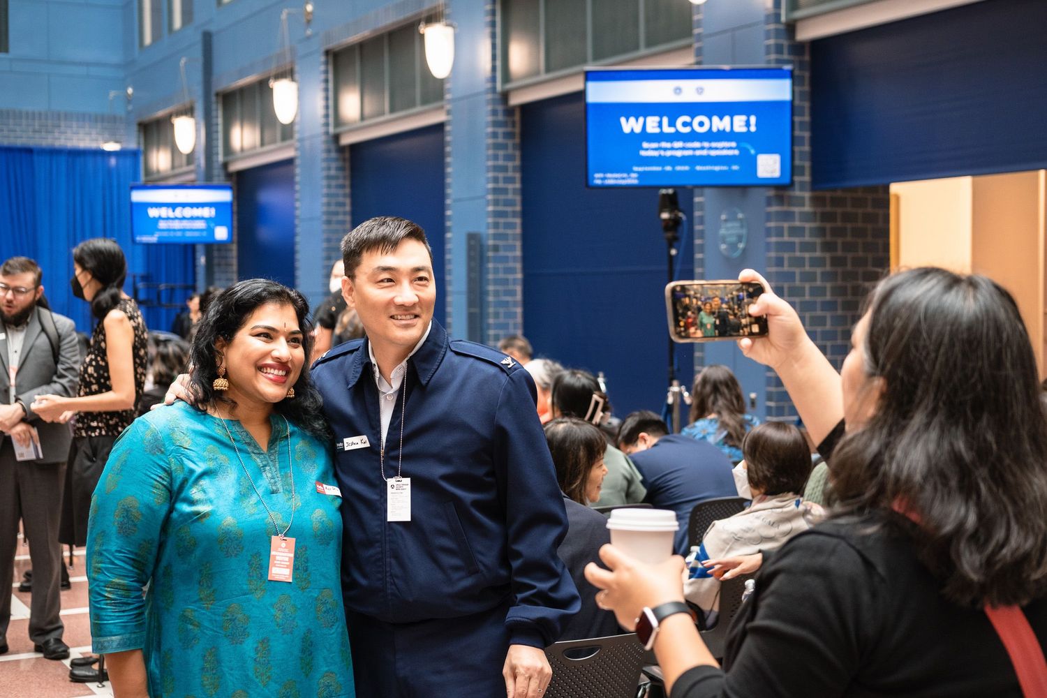 Two professionals pose for a photo at a welcome event, with a blue WELCOME sign visible in the background.