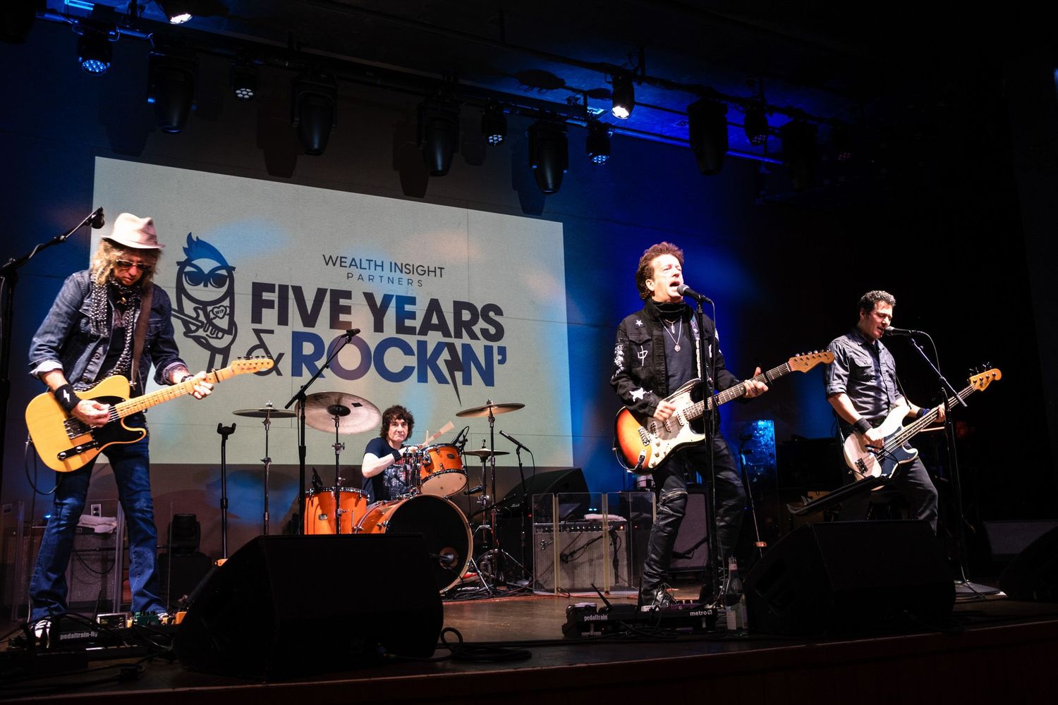 Four musicians performing on stage at Wealth Insight Five Years Rockin anniversary event with guitars and drums.