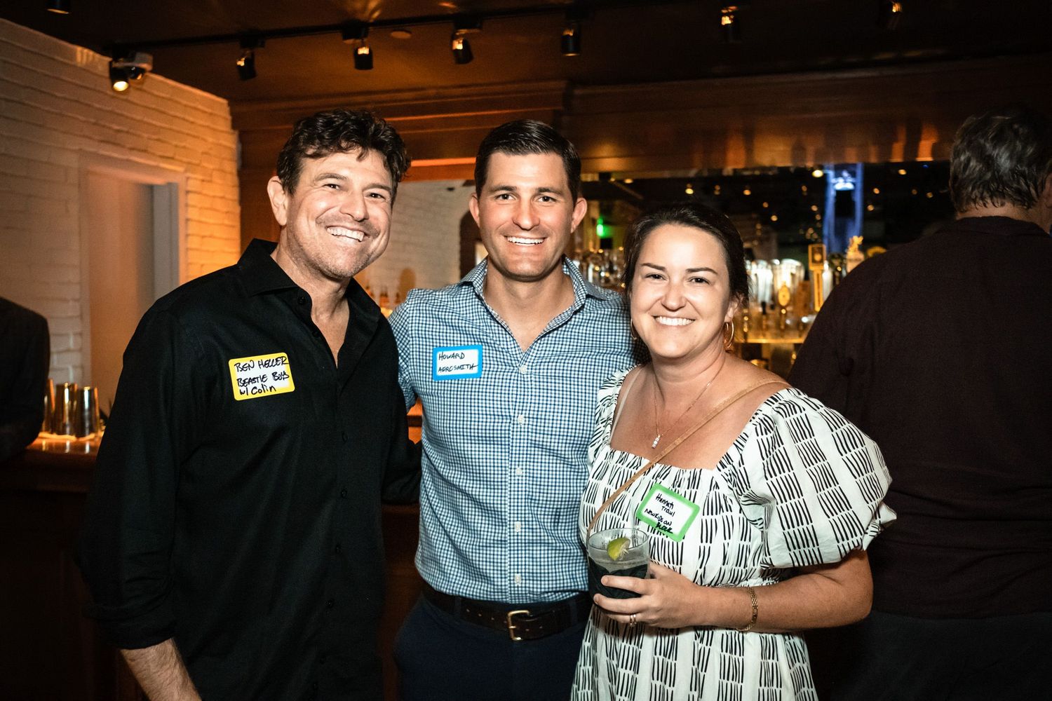 Three smiling friends pose together at an indoor networking event, with name tags and drinks in hand.