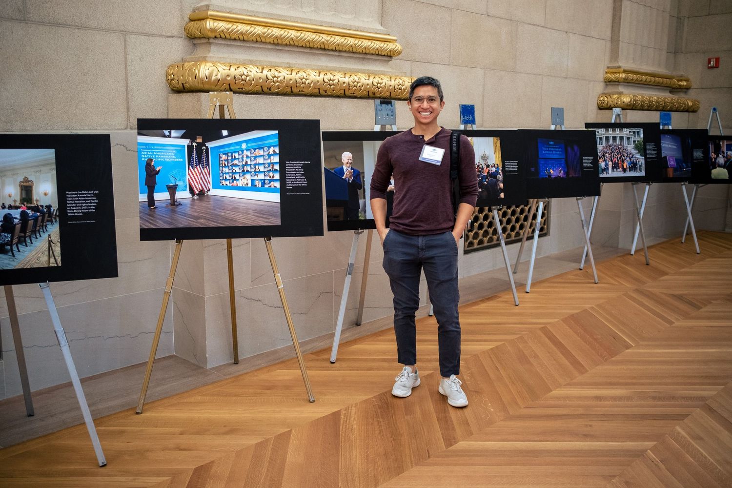 Young man smiling at photo exhibition display on wooden parquet floor in elegant gold-accented gallery hall.