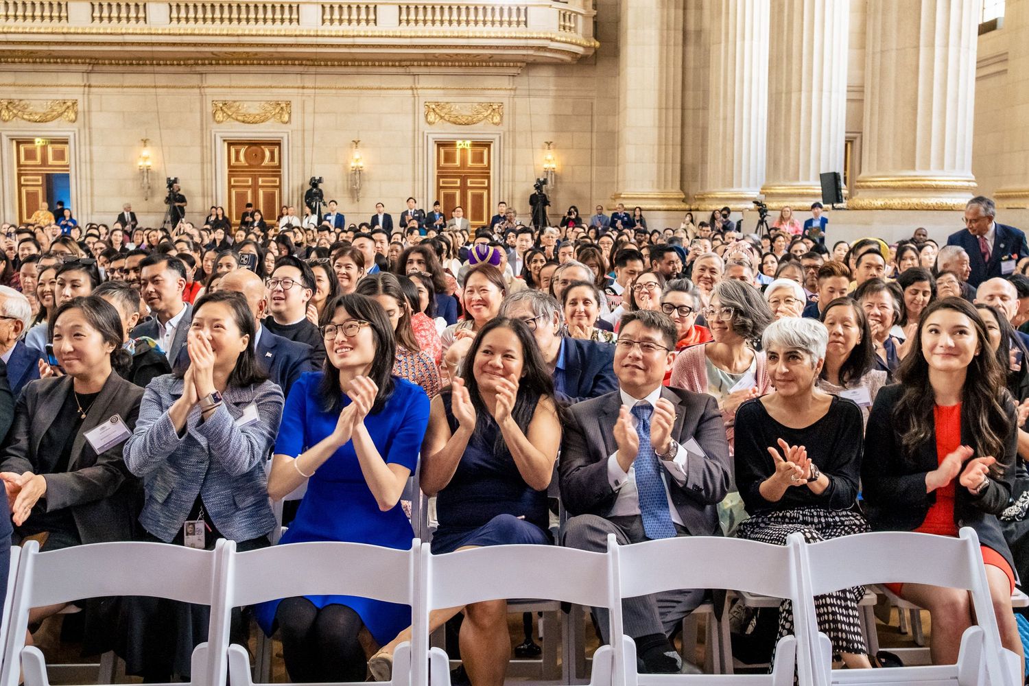 Diverse crowd of people applauding and smiling at a formal event inside an elegant government building.