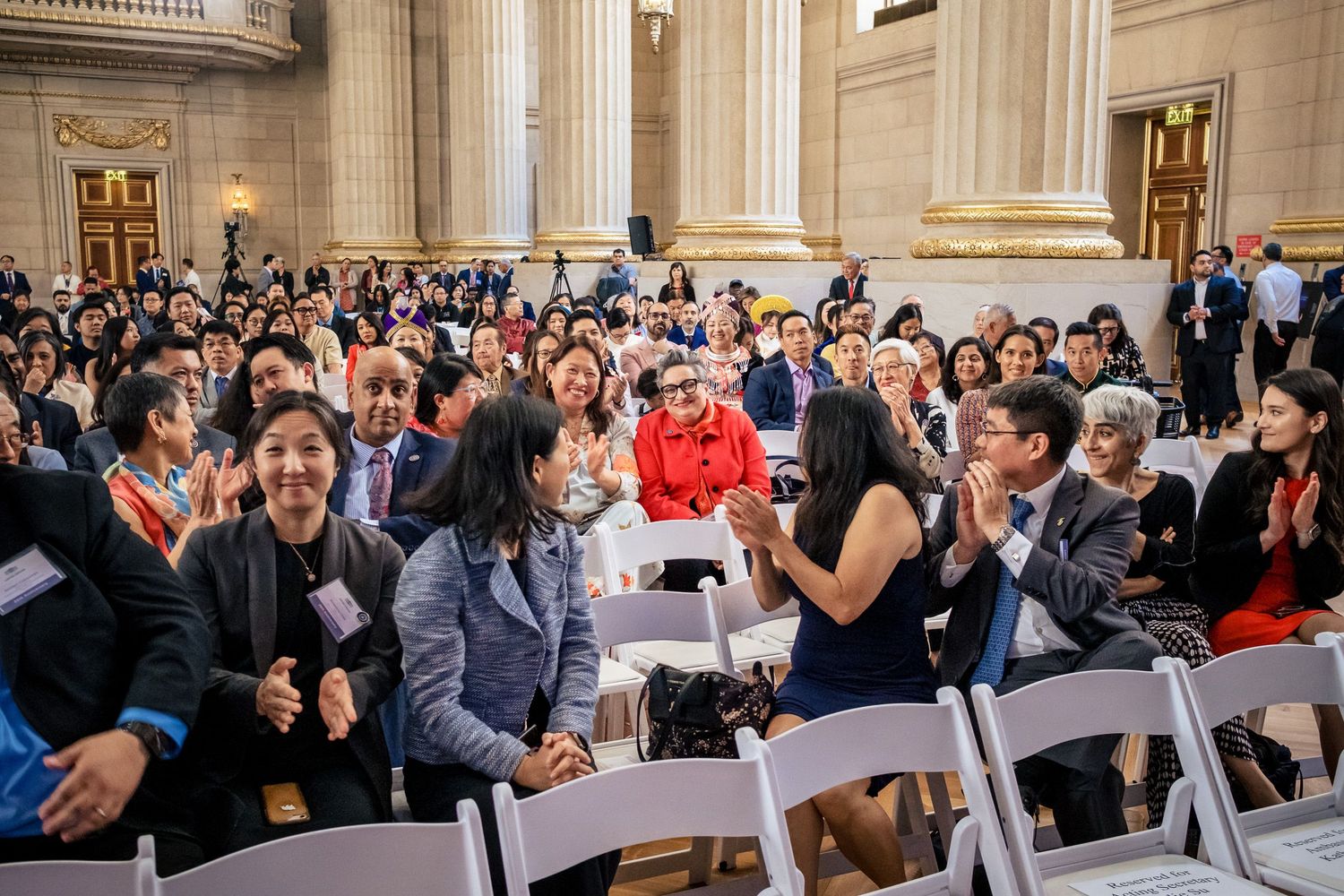 Diverse crowd applauding at an indoor ceremony in a grand hall with columns.