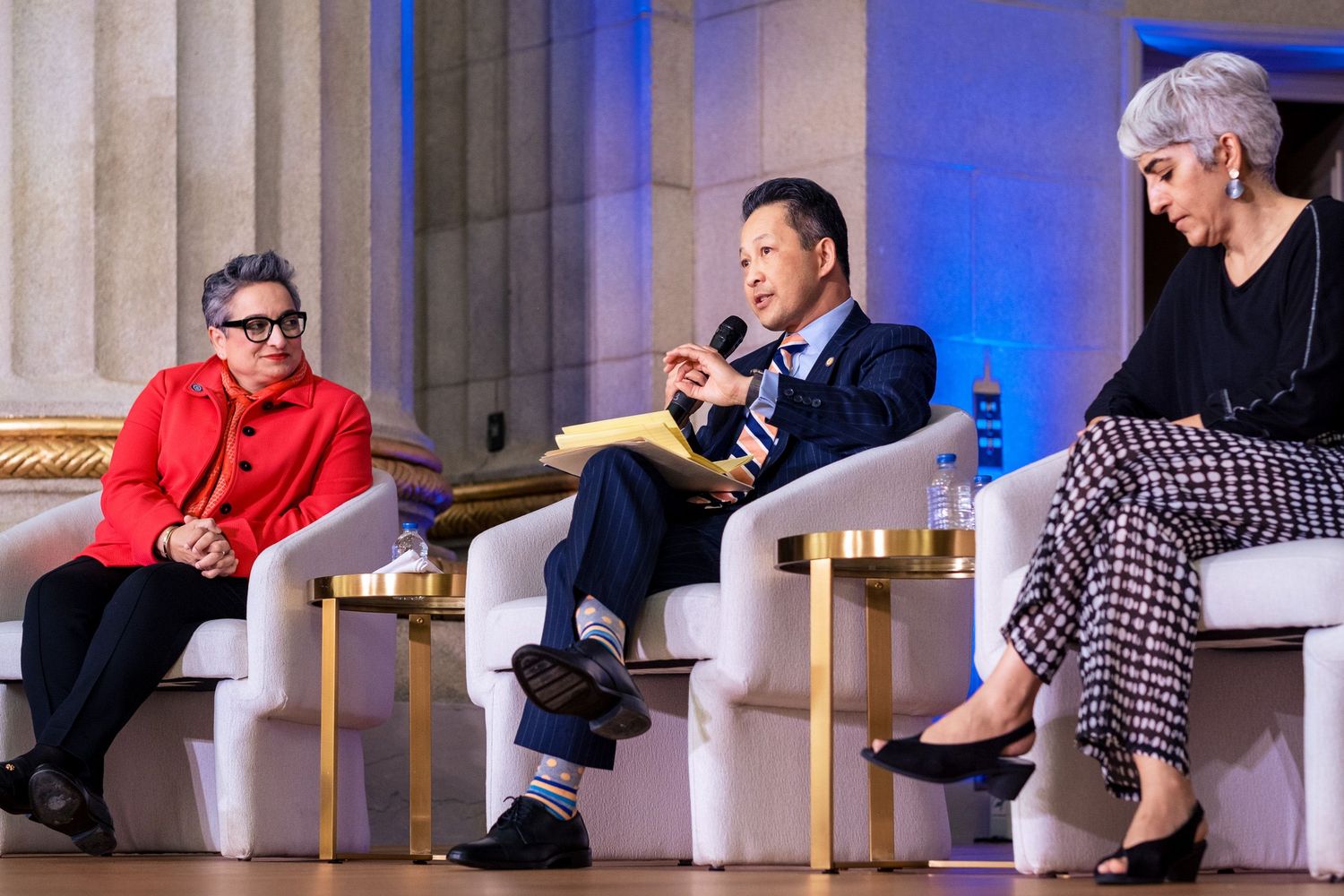 Three panelists engaged in discussion on stage at a formal conference event, seated in white chairs.