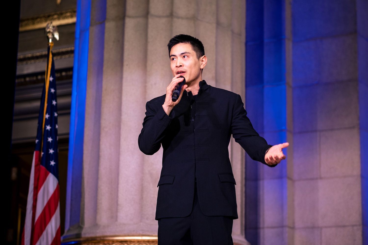 Young Asian man in black suit speaking into microphone on stage with American flag and columns behind him.
