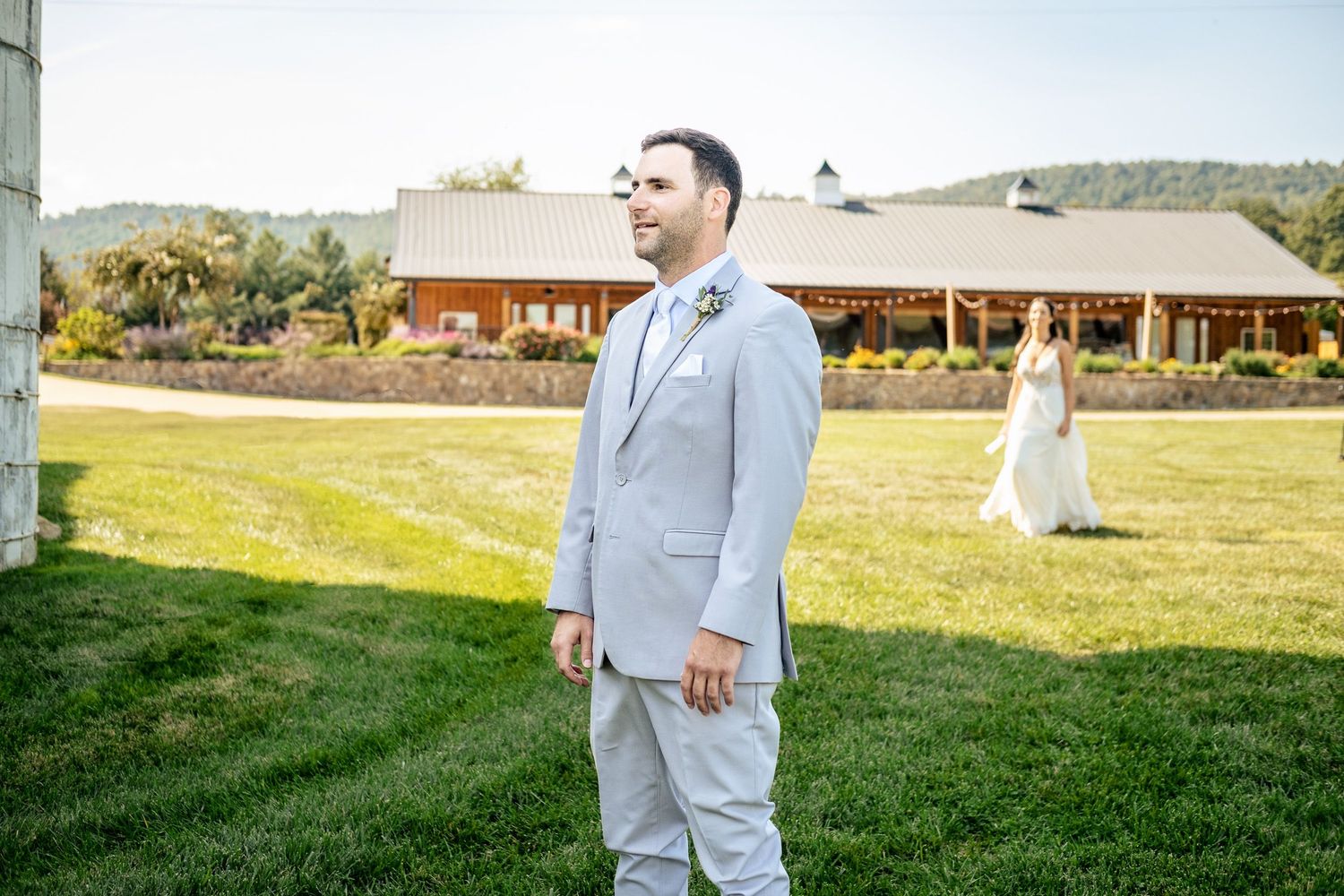 Groom in light gray suit stands on green lawn as bride in white gown approaches from rustic barn venue.