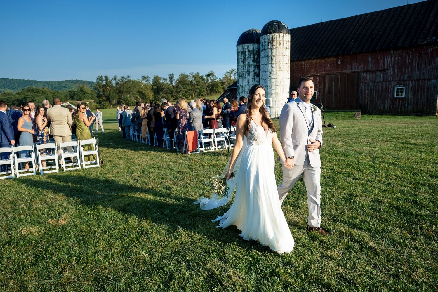 Newlyweds walk hand in hand across green grass after outdoor barn wedding ceremony with guests seated behind.