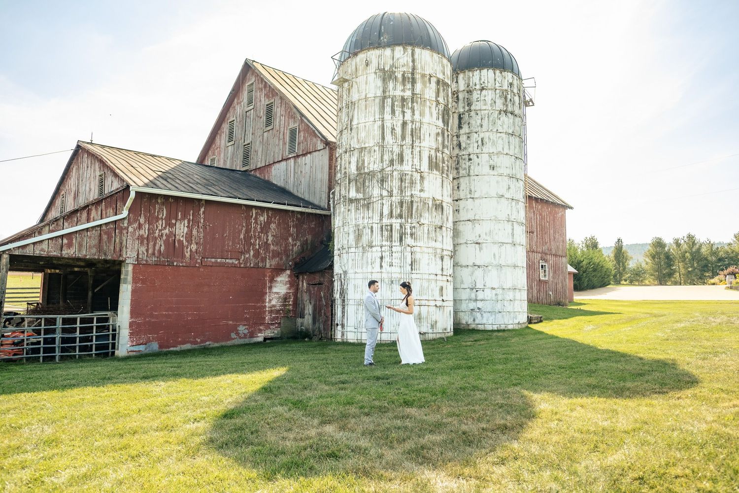 Bride and groom dance joyfully in front of classic red barn with twin white silos.