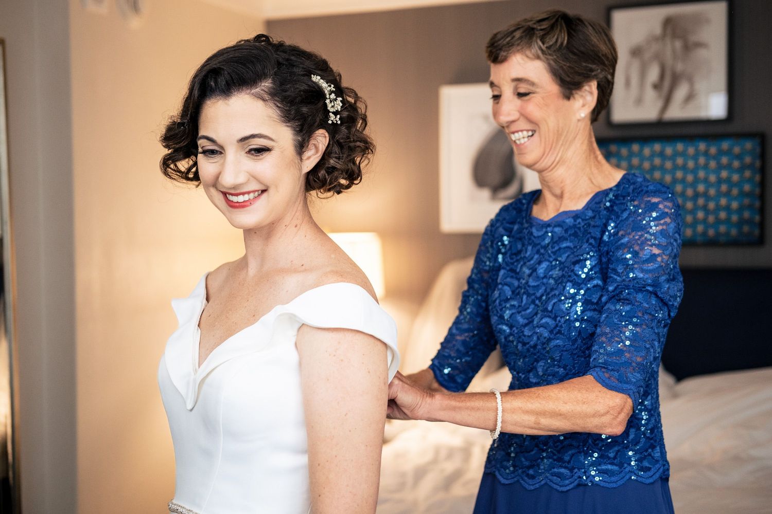 Smiling bride in white off-shoulder gown gets dress fastened by woman in blue lace dress.