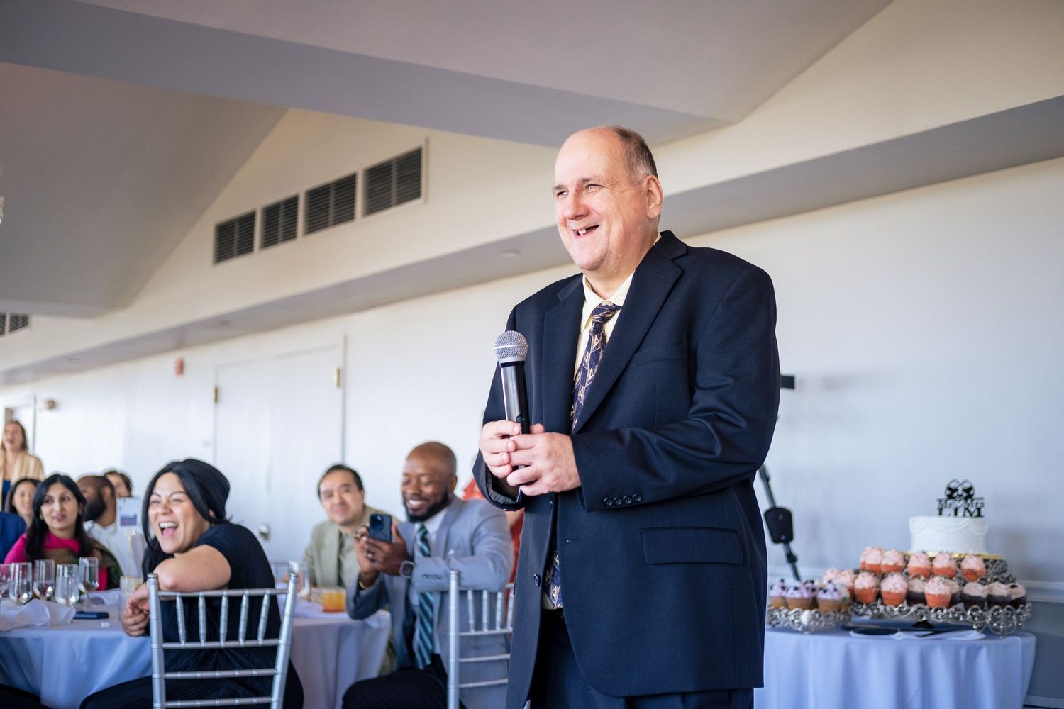 Man in suit smiles while giving a wedding speech to guests seated at decorated reception tables.