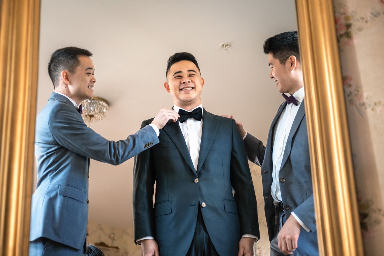 Two groomsmen helping groom adjust tuxedo bow tie in elegant gold mirror.