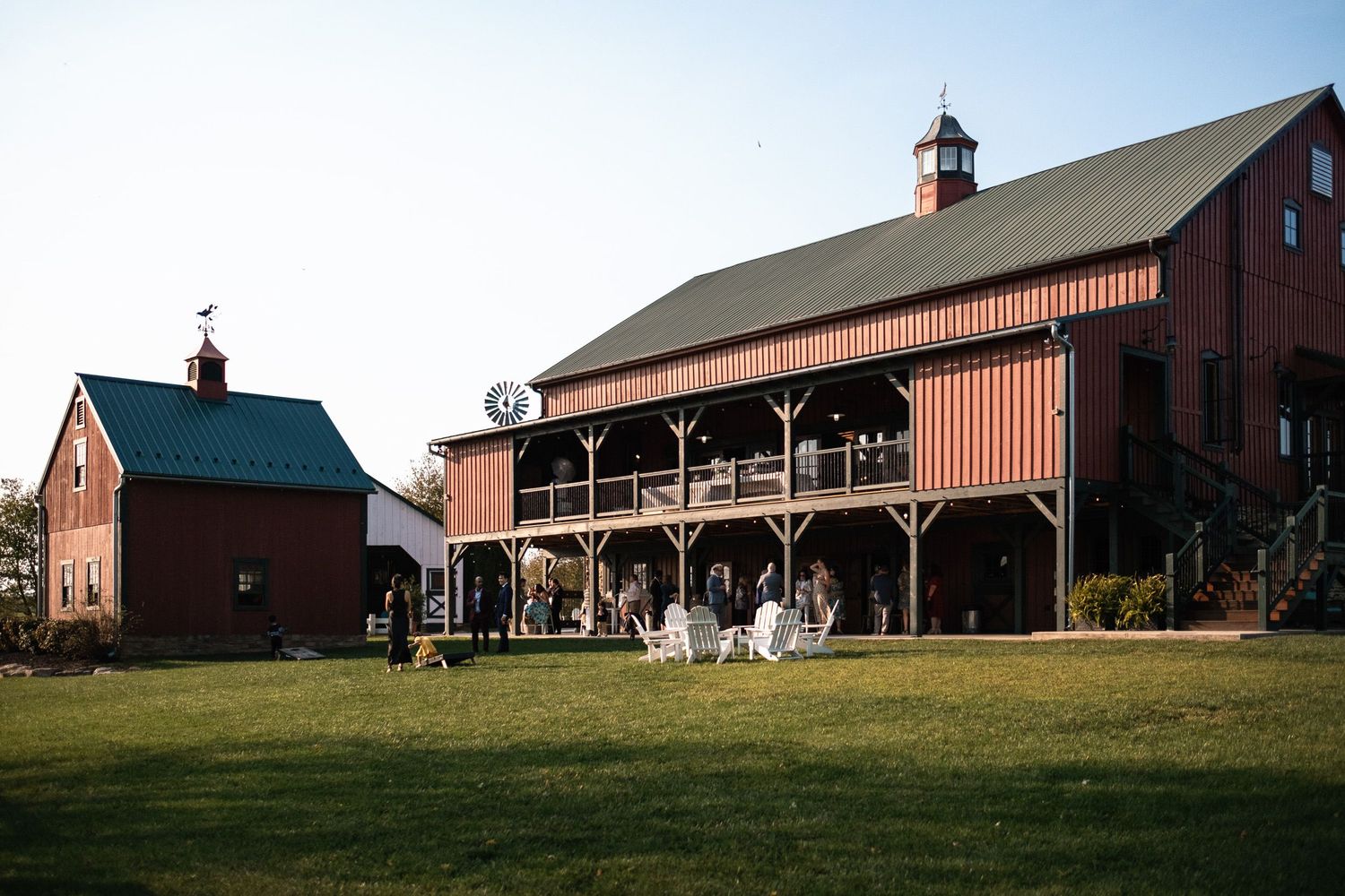 Red barn wedding venue exterior with large deck and guests gathered on green lawn at sunset.