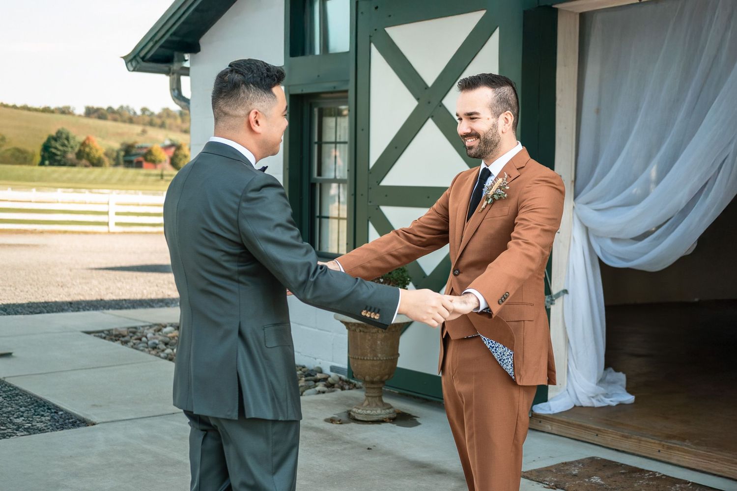 Two grooms in suits shake hands during an emotional first look at a barn wedding venue.