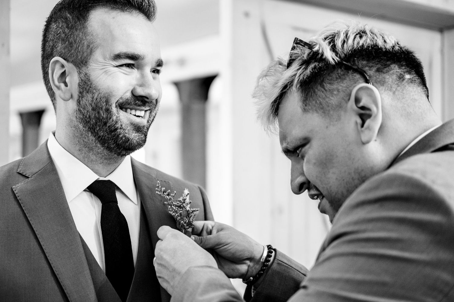 Black and white photo of smiling groom having boutonniere pinned on lapel by groomsman.