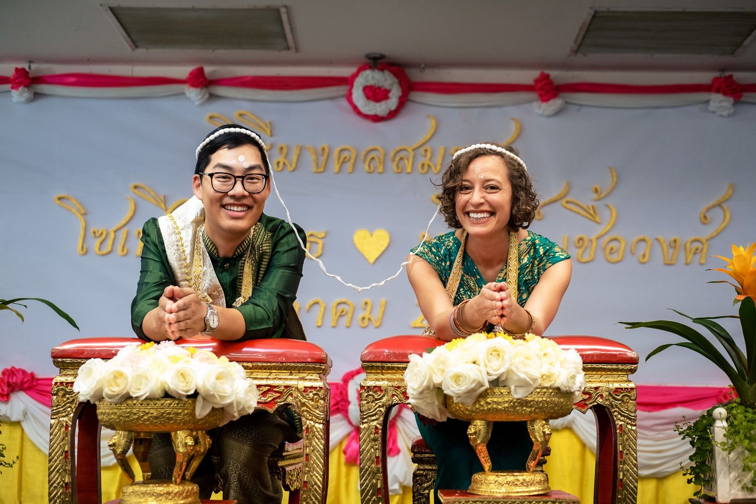 Couple in traditional Thai wedding attire smiling at ceremony with golden decorations and floral arrangements.