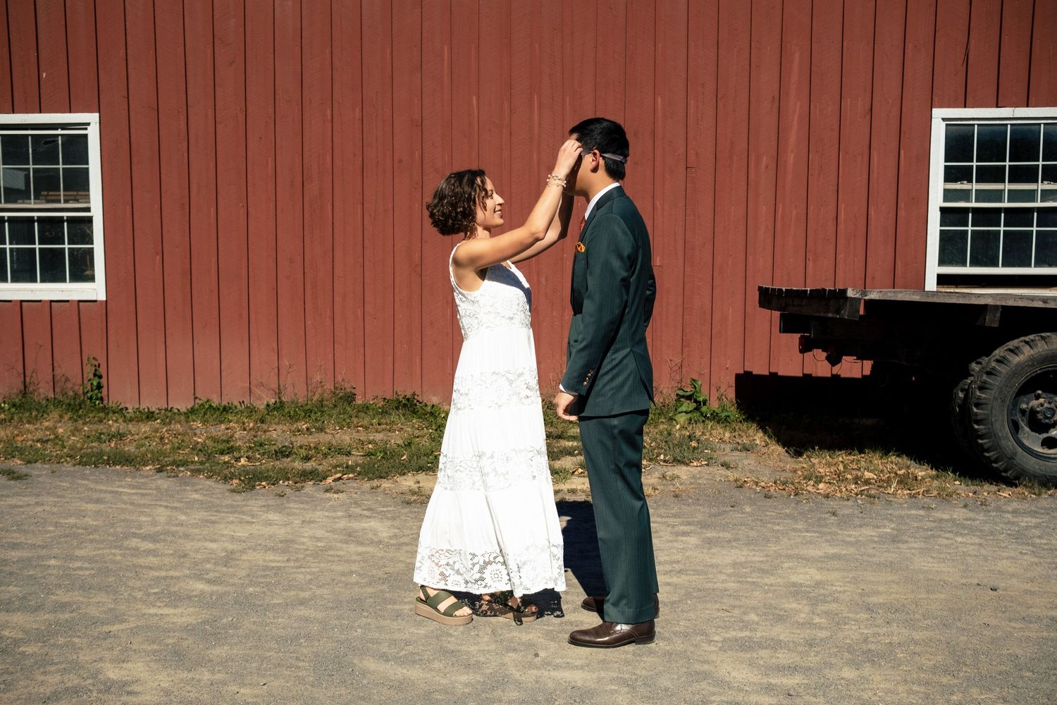 Bride removing blindfold from groom during first look moment beside red barn.