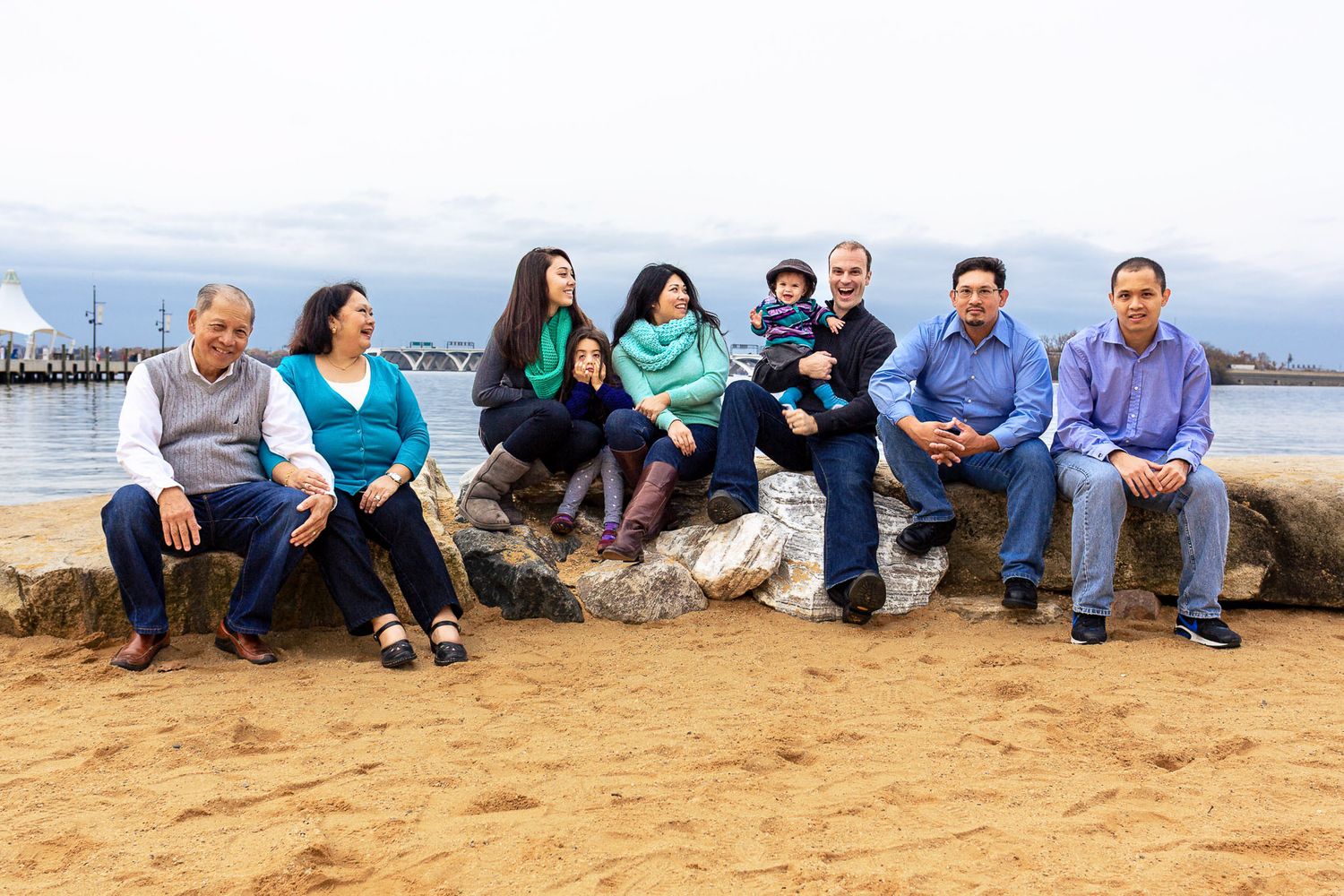 Extended family group photo sitting on rocks at a sandy beach with water in background.