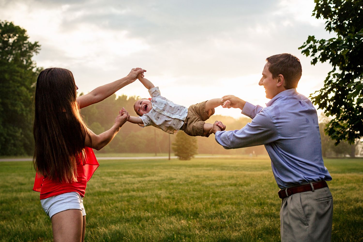 Happy couple swinging their young child between them in a grassy park at golden hour sunset.