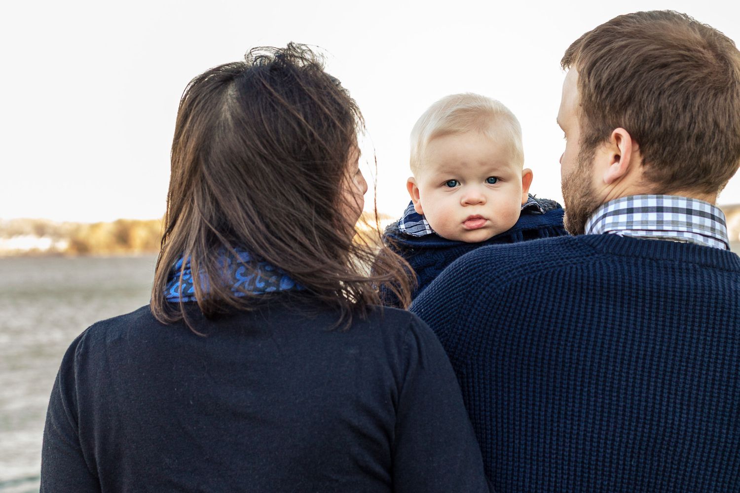 Parents holding their curious baby boy while standing together near a serene waterfront.