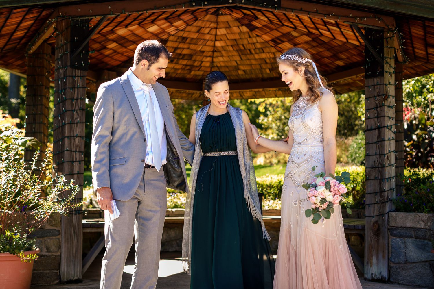 Bride in beaded gown meets officiant and guest under wooden gazebo in sunny garden setting.