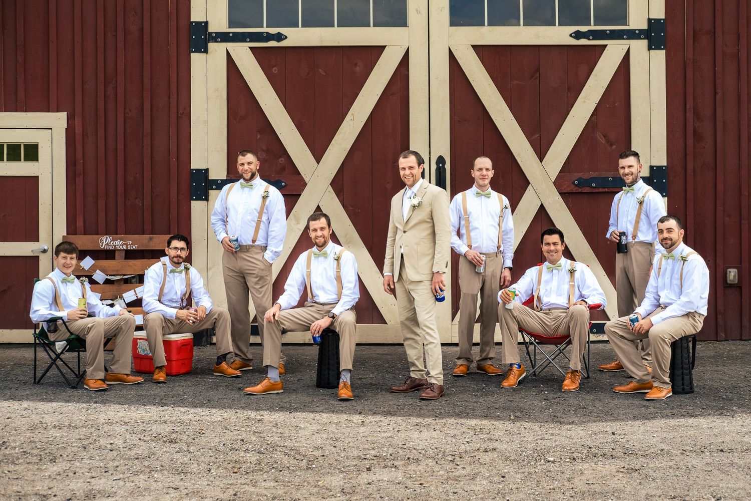 Groom and groomsmen pose casually in tan suits with suspenders outside a red barn.