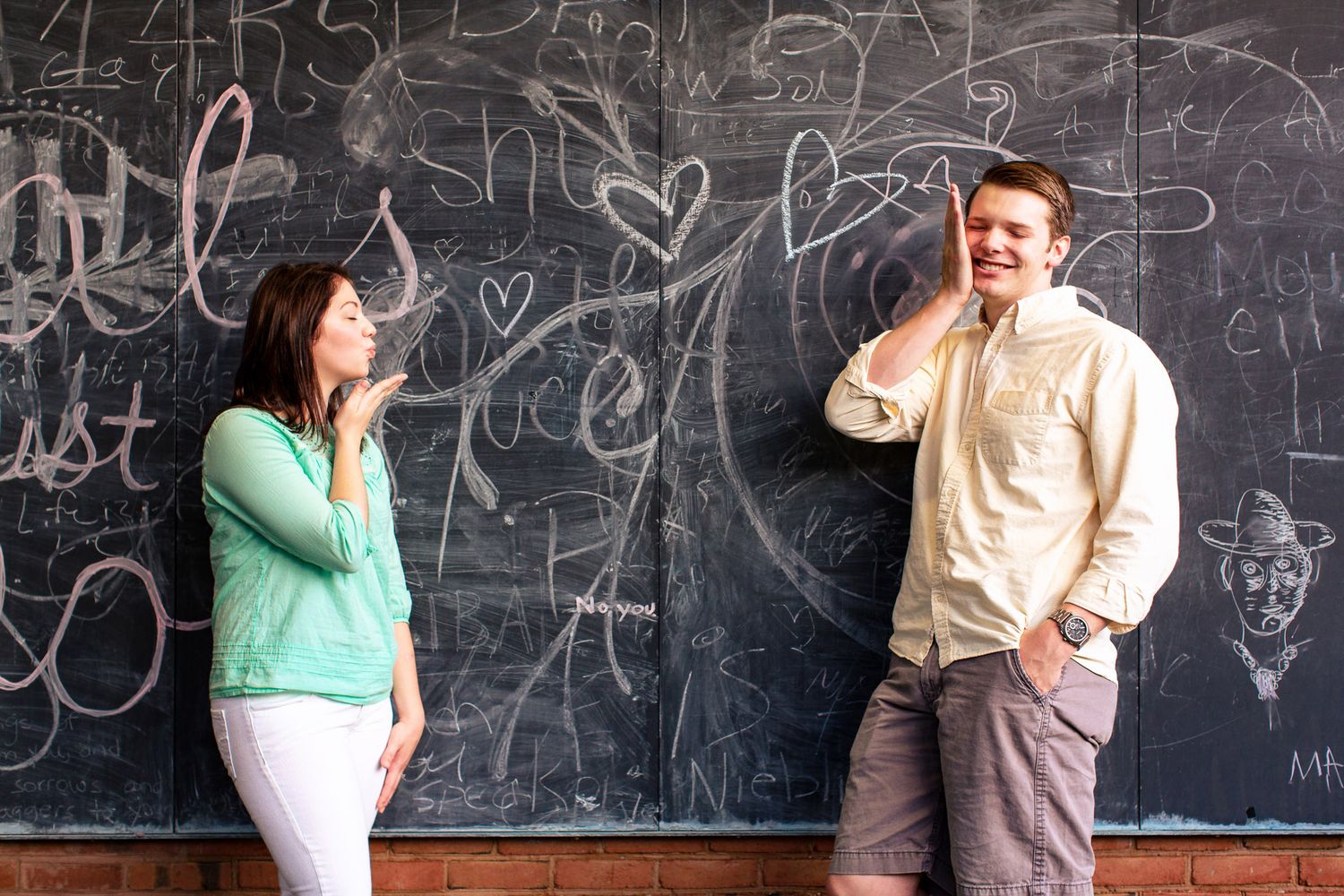 Man and woman laughing together in front of a chalkboard covered in hearts and doodles.