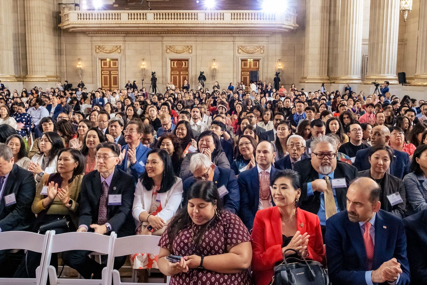 Diverse crowd standing and applauding at a formal event inside a grand neoclassical building.