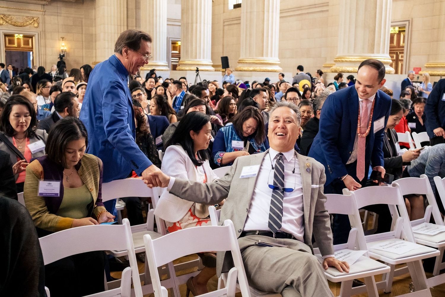 Man laughing in chair at crowded indoor event while others interact around him in ornate hall.