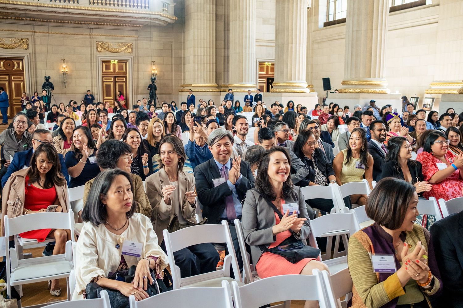 Large diverse crowd of smiling attendees applauding at an indoor event in a grand hall with columns.
