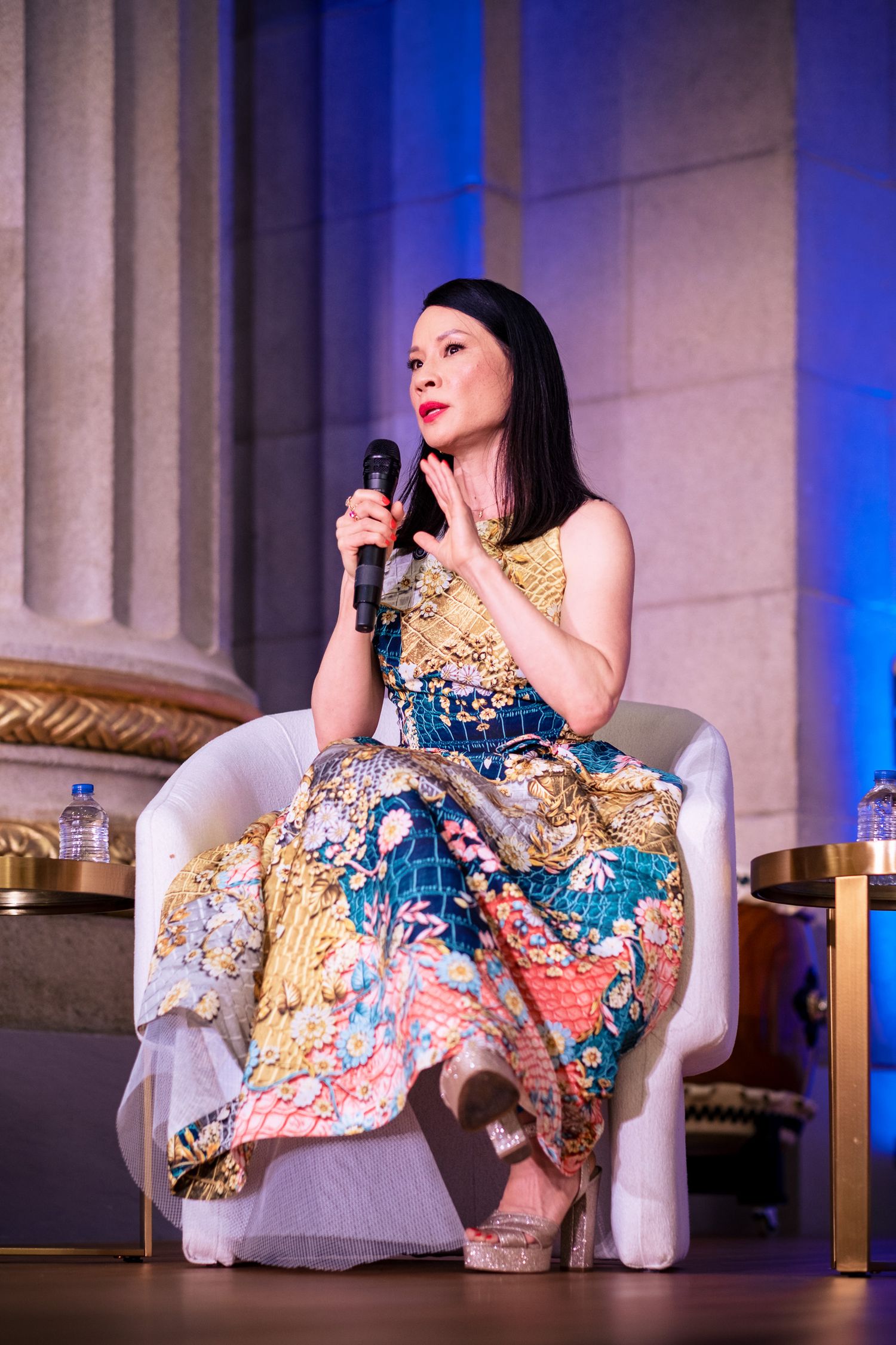 Woman in colorful floral dress speaks into microphone while seated on white chair at elegant event.