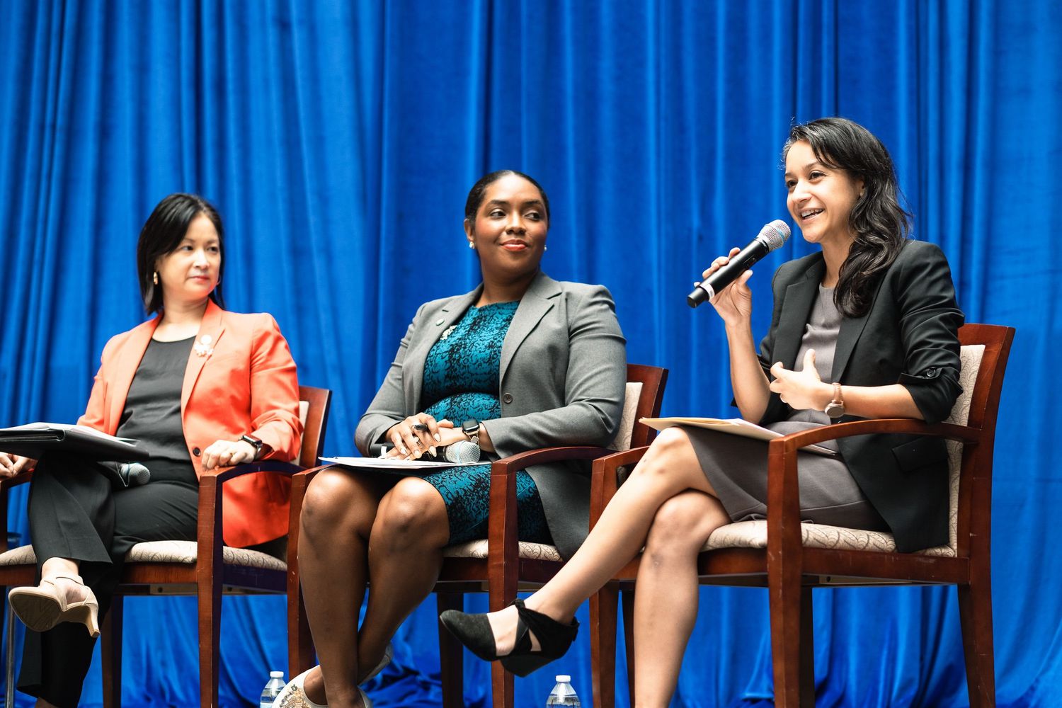 Three professional women seated on stage at a panel discussion, one speaking into a microphone before a blue curtain backdrop.