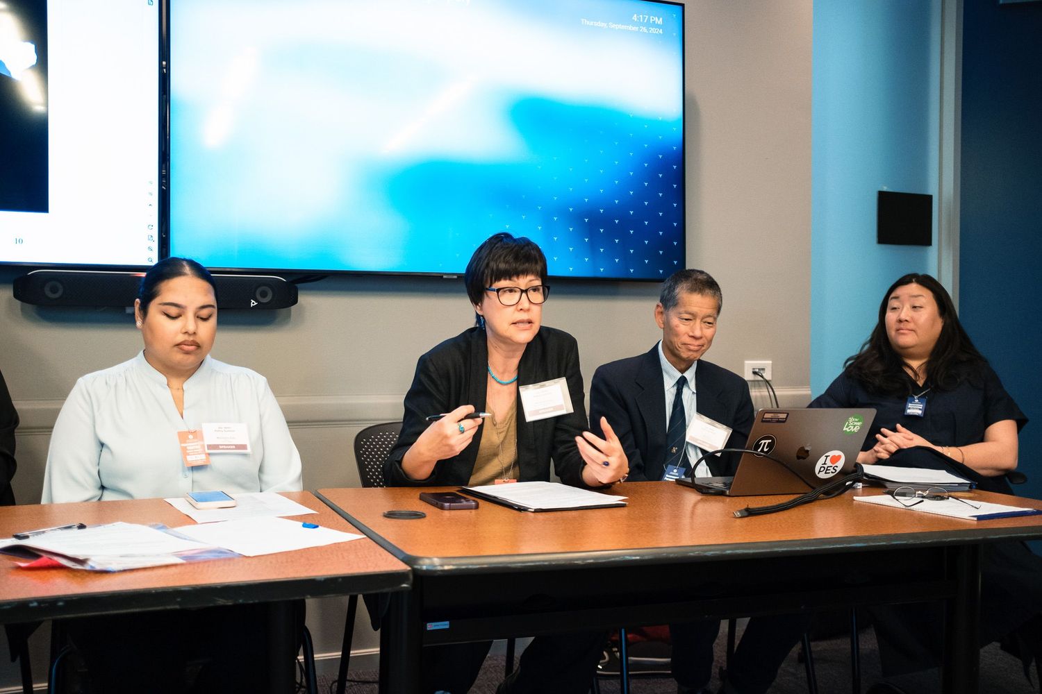Four professionals sit at a conference table during a panel discussion with a screen displayed behind them.