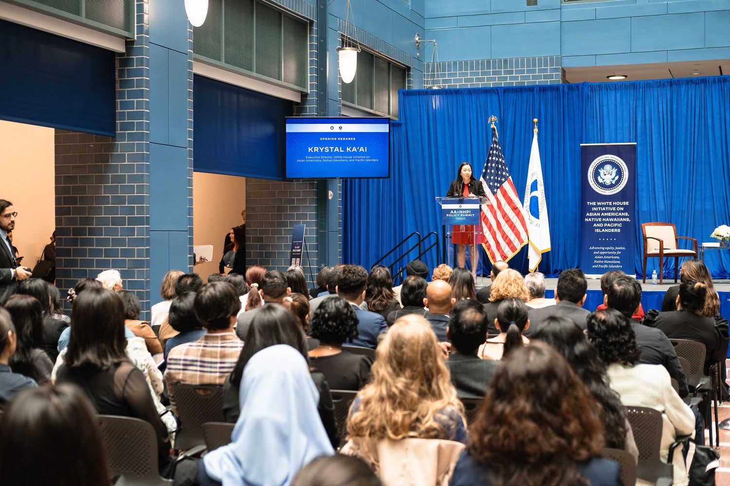 Speaker addresses a diverse audience at a formal government event with US and agency flags displayed on stage.