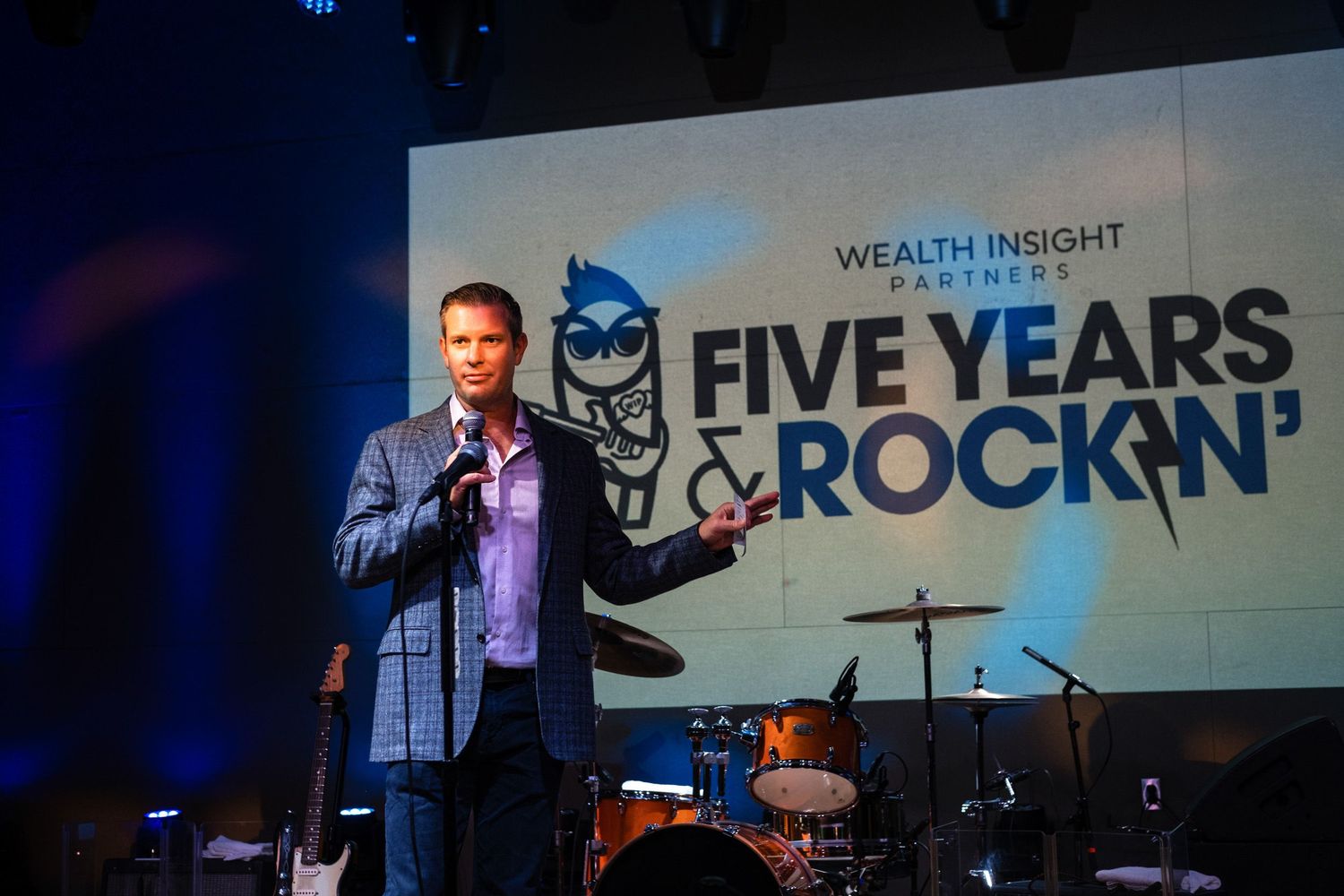 Man speaking on stage at Wealth Insight Partners Five Years Rockin anniversary event with band equipment visible.