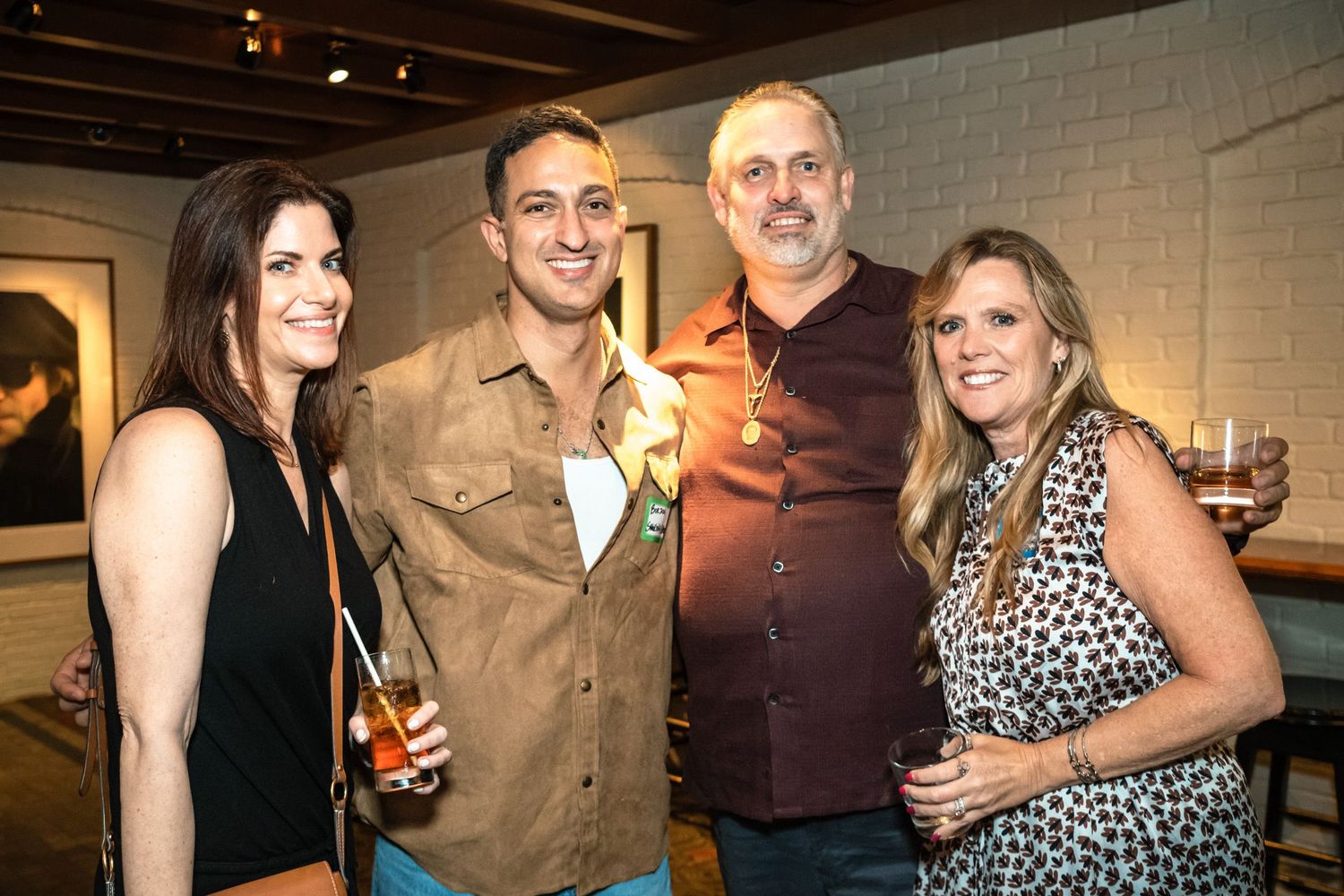 Four friends smiling and holding drinks at a casual indoor social gathering or party event.