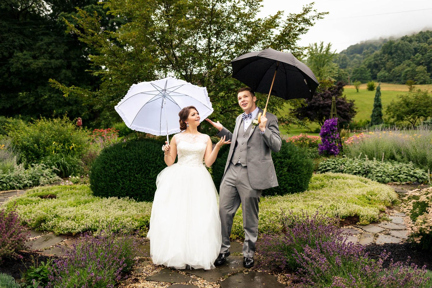 Bride and groom pose playfully with umbrellas in a lush garden on a rainy wedding day.