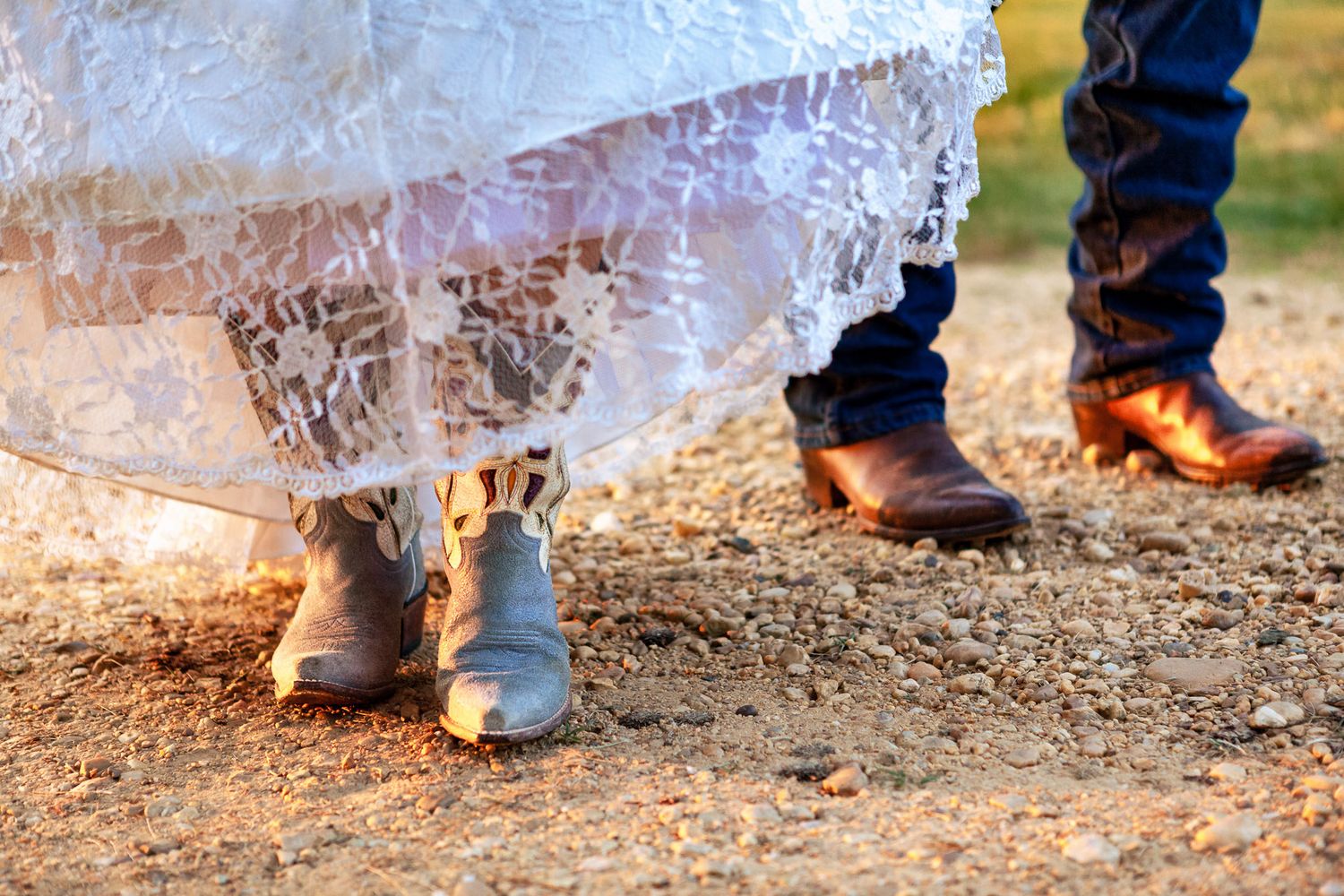 Bride and groom wearing cowboy boots beneath a lace wedding dress on rustic ground.