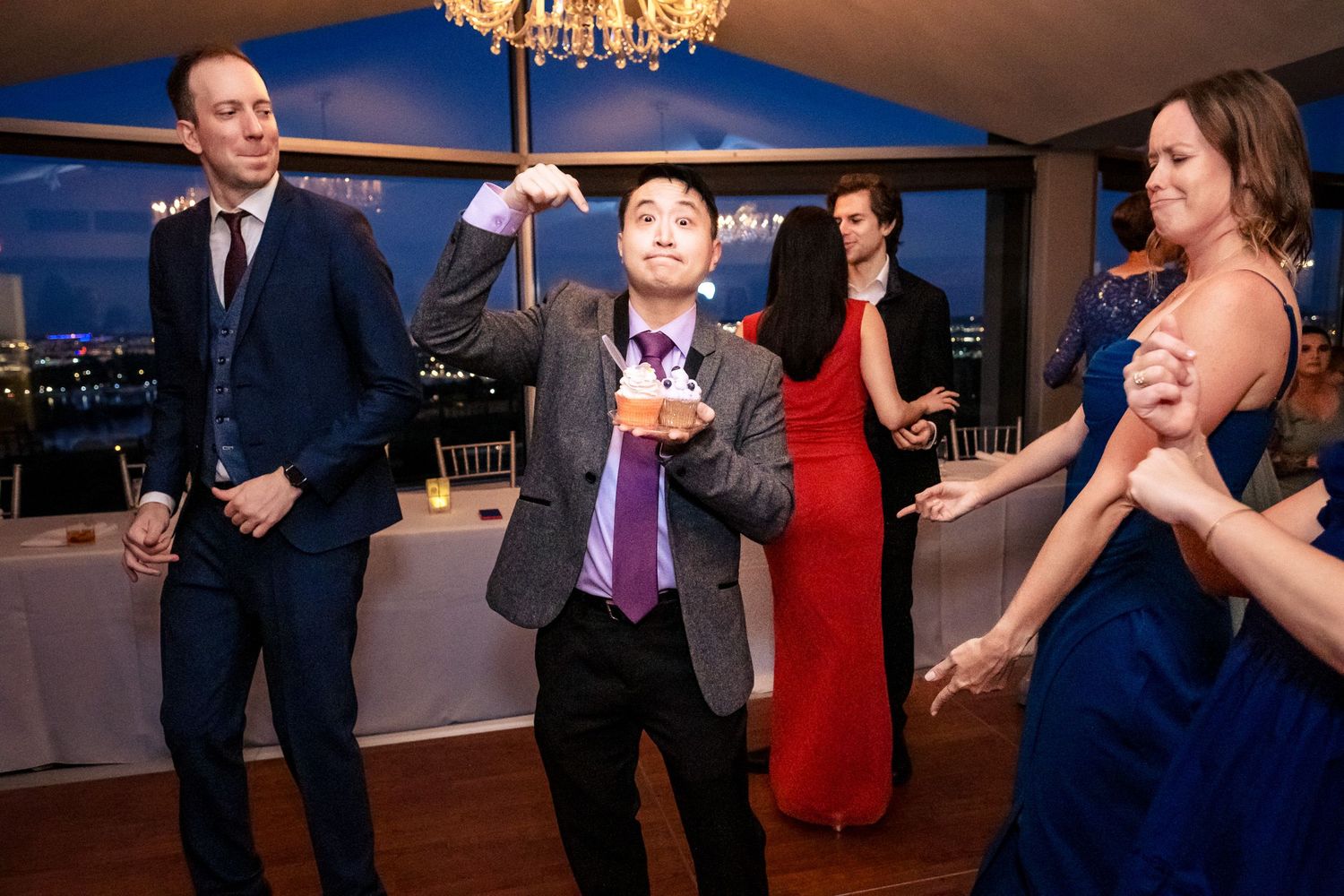 Wedding guest dances energetically holding a small cake at an elegant indoor reception venue.