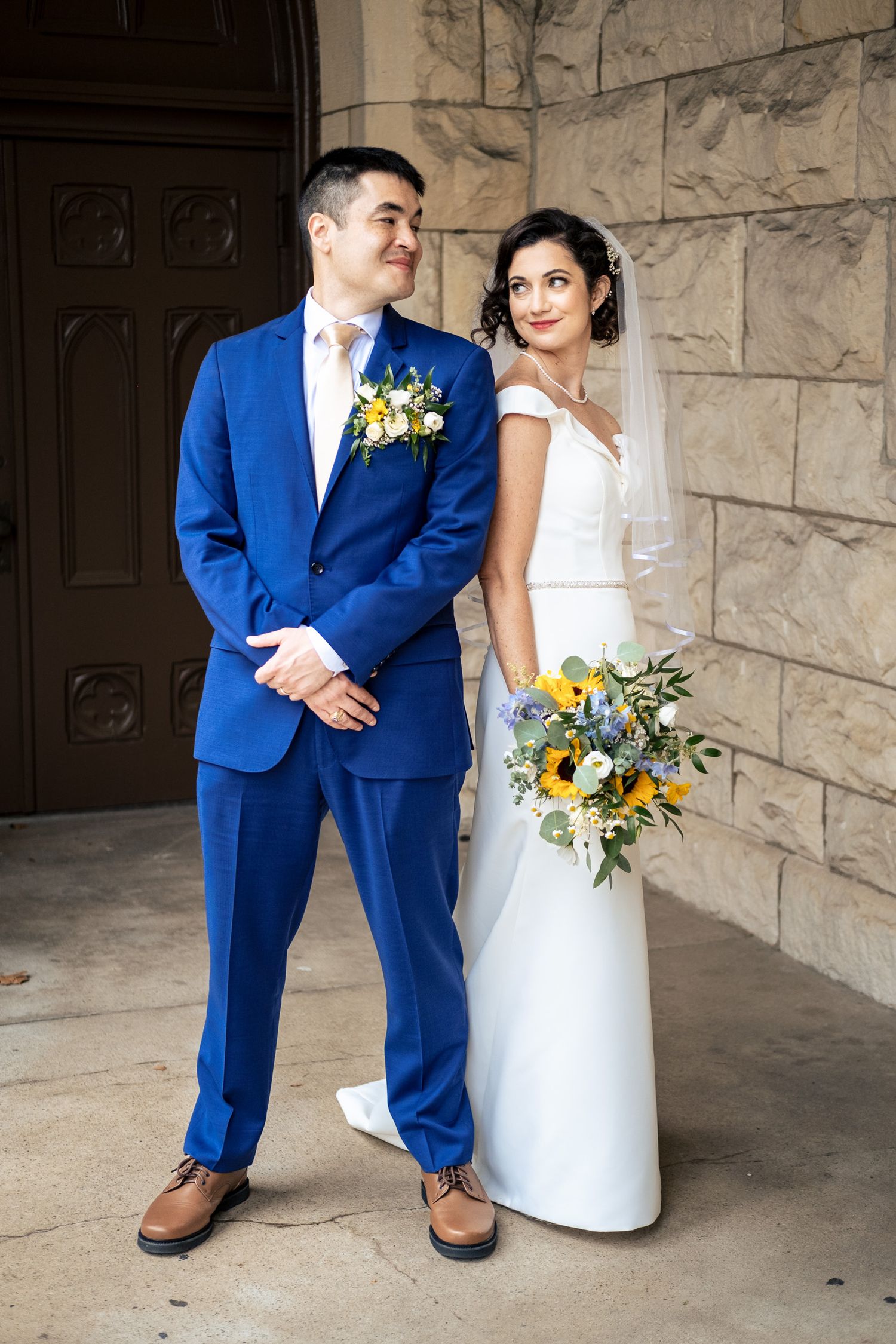 Bride in white gown with veil and groom in royal blue suit pose together outside a stone church.