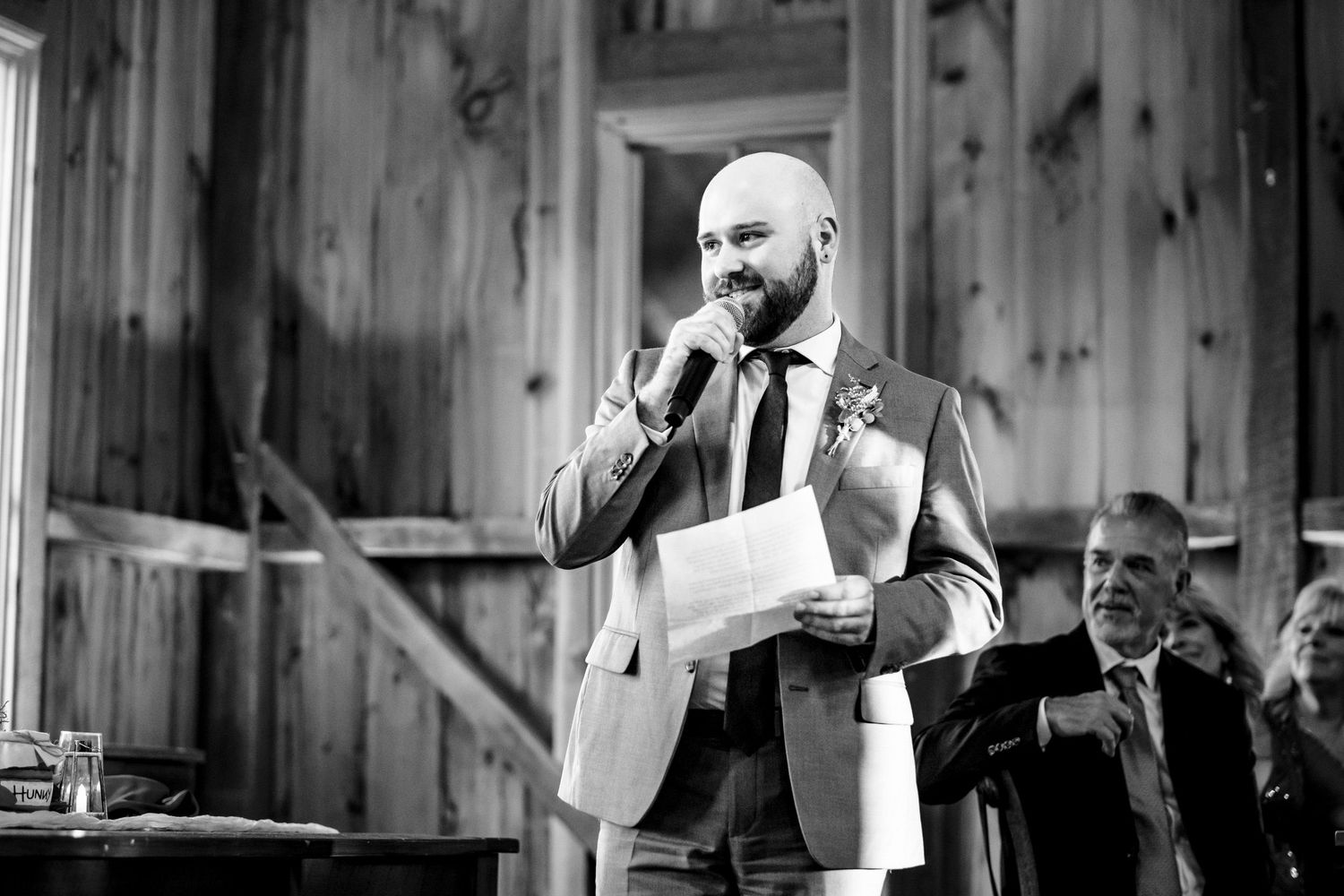 Bearded man in suit reading wedding speech from paper while holding microphone in barn venue.