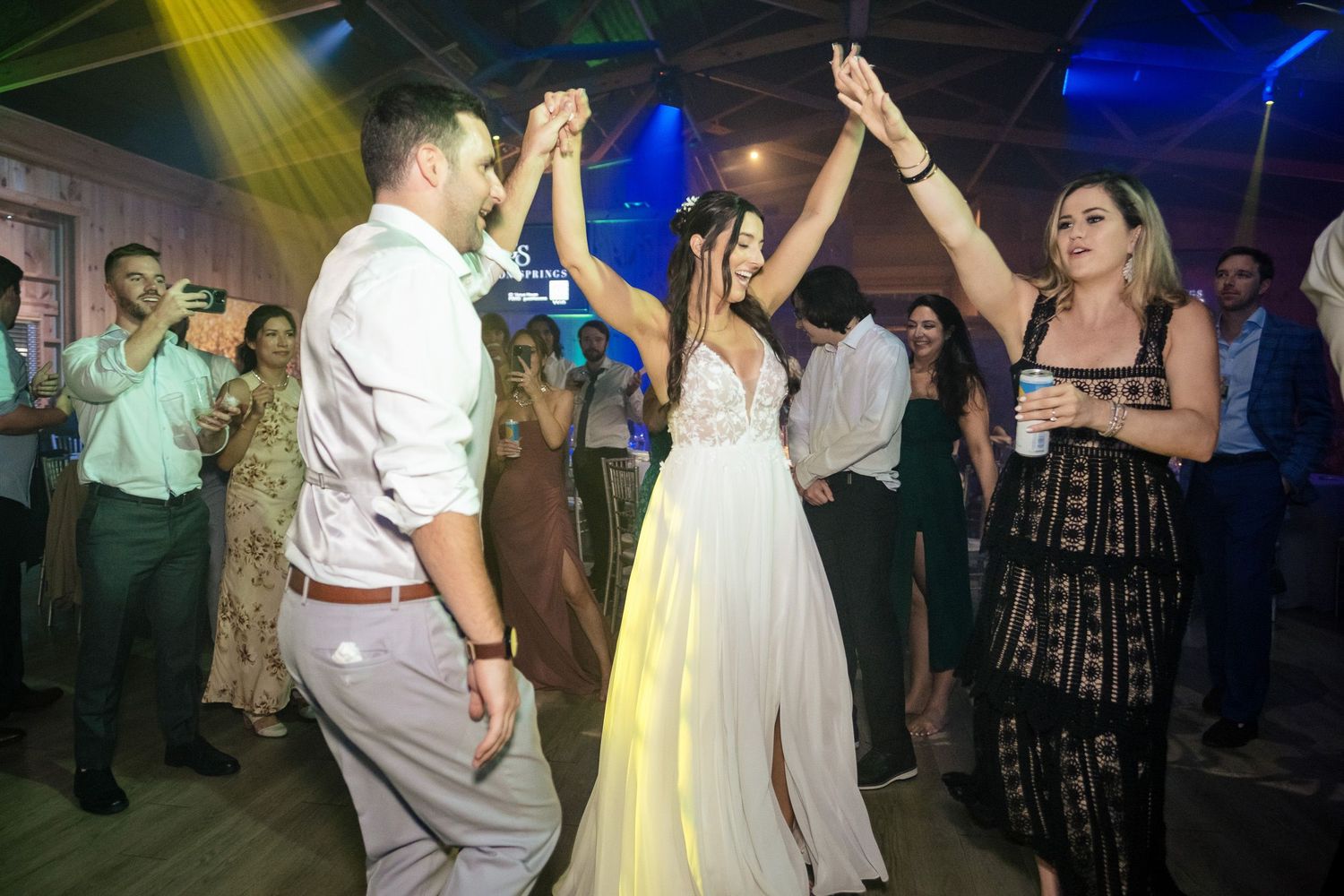 Bride in white gown dancing with groom and guest under colorful lights at wedding reception.