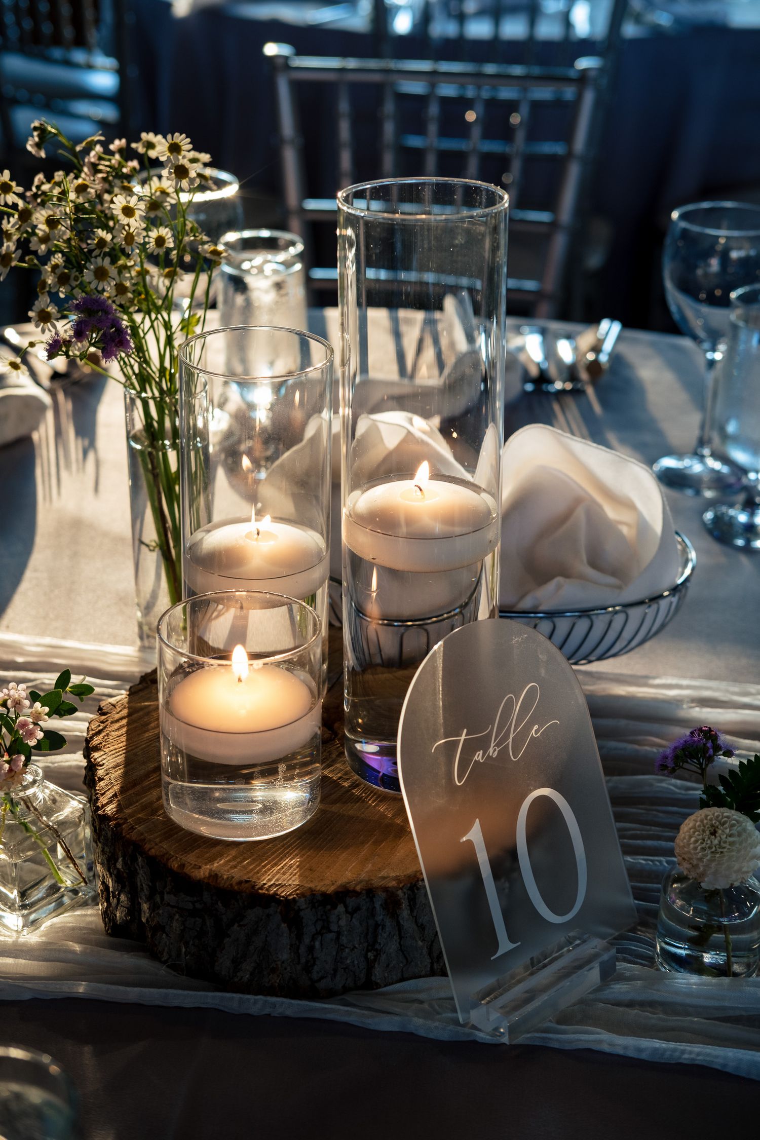 Elegant wedding table centerpiece featuring floating candles in glass cylinders on a wood slice with wildflowers.