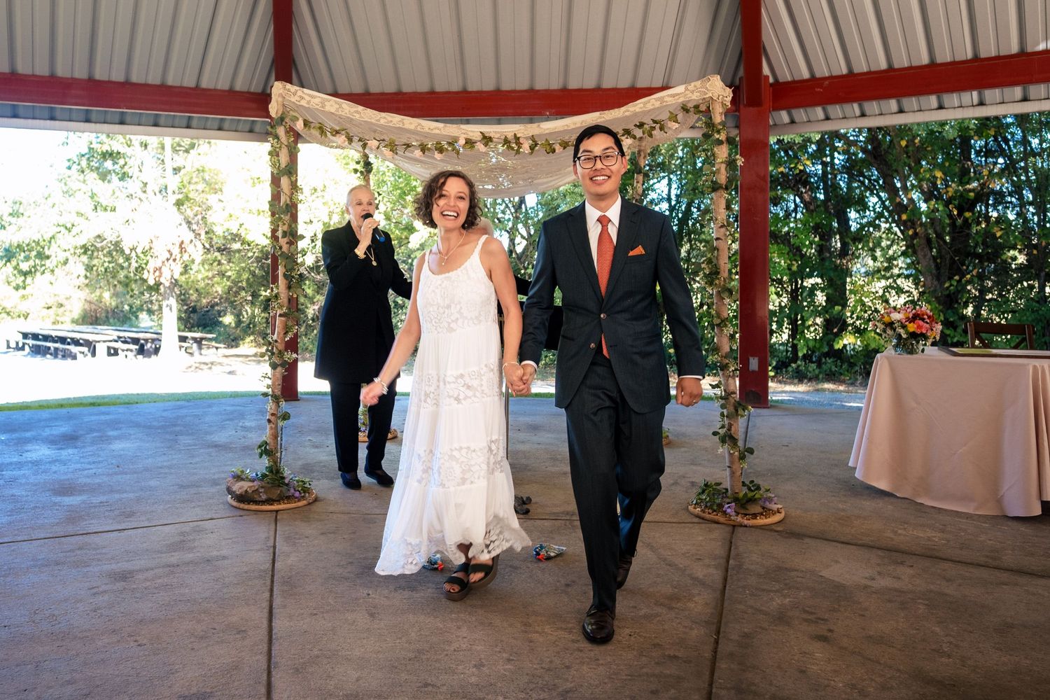 Newlyweds walk hand in hand after outdoor wedding ceremony under floral chuppah gazebo.