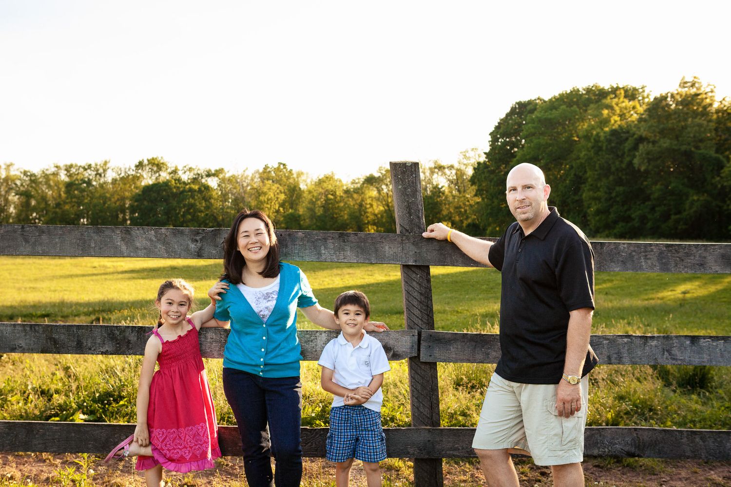 Family of four smiling together beside a wooden fence post in a sunny green field at golden hour.